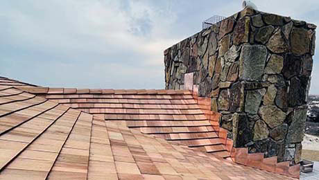 A rooftop with new reddish-brown wooden shingles slopes toward a large stone chimney under a partly cloudy sky, expertly installed by a roofer Long Island trusts.