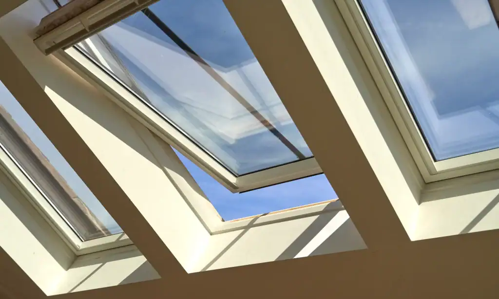 A row of rectangular skylight windows in a ceiling, expertly installed by a roofing contractor Long Island, with one window partially open, letting in natural sunlight and revealing a clear blue sky outside.