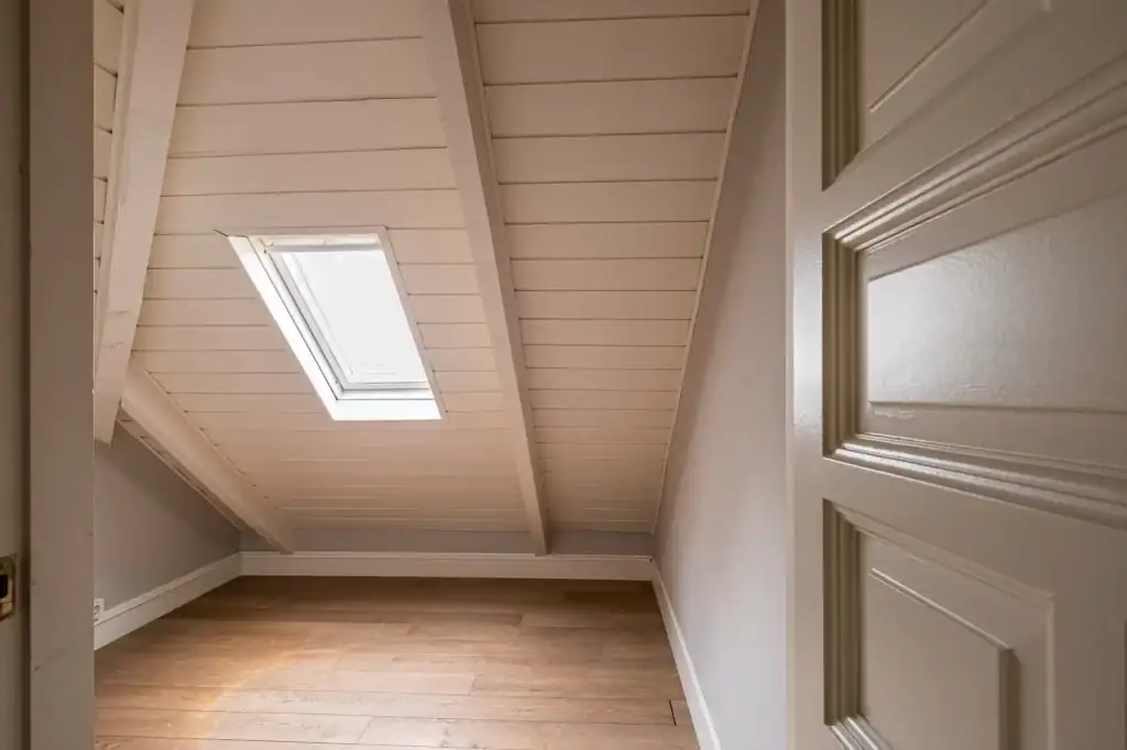 A small attic room with a slanted white wooden ceiling, installed by a skilled roofing contractor Long Island, light gray walls, and wooden flooring. The image is taken from the doorway, capturing the cozy, empty space.