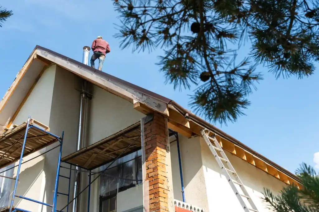 A person in work clothes stands on the roof of a house under construction, with scaffolding, a ladder, and trees visible—showcasing the expertise of a roofer Long Island under a clear NY blue sky.