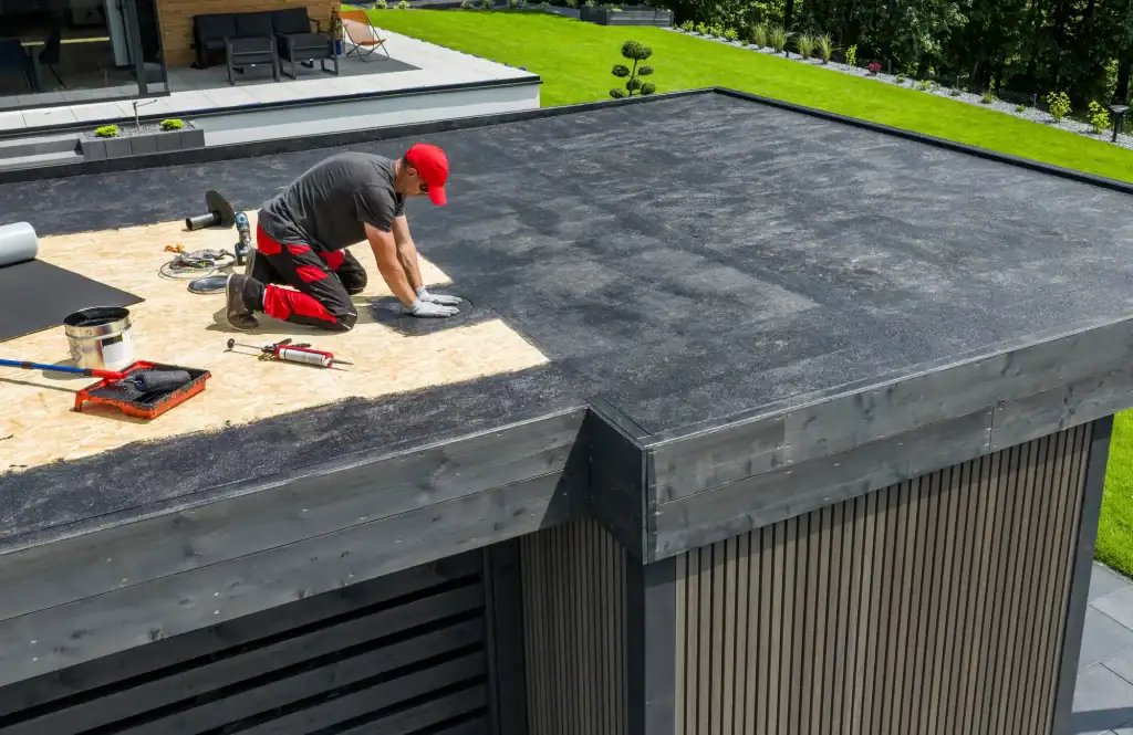 A roofer Long Island in a red cap and gloves kneels on a flat rooftop, installing roofing material with tools and supplies nearby. The roof overlooks a green lawn and landscaped garden.