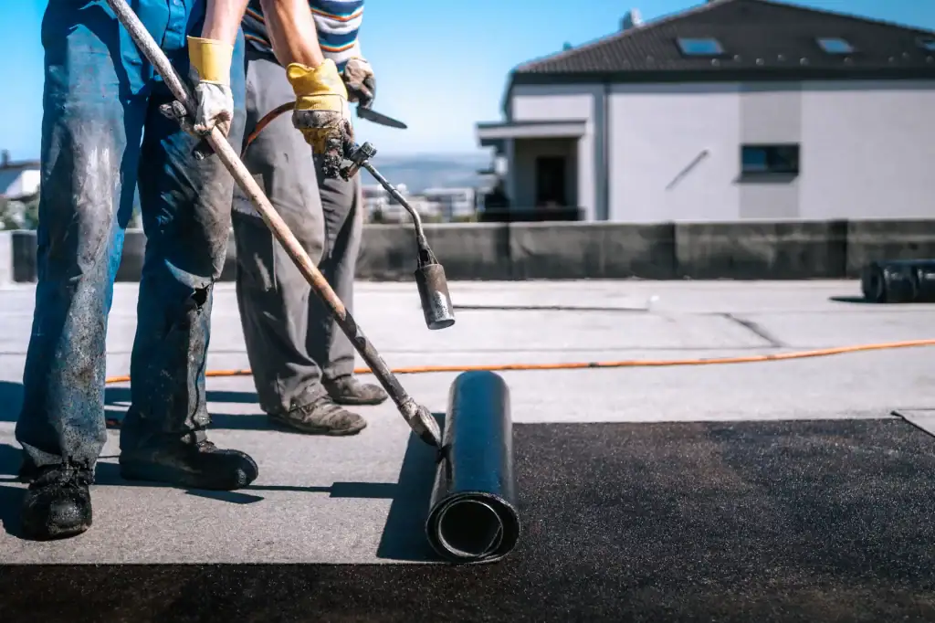 Two workers in gloves and work clothes, from a roofing contractor Long Island, use a torch to install and seal a roll of roofing material on a flat rooftop under a clear sky, with a building visible in the background.