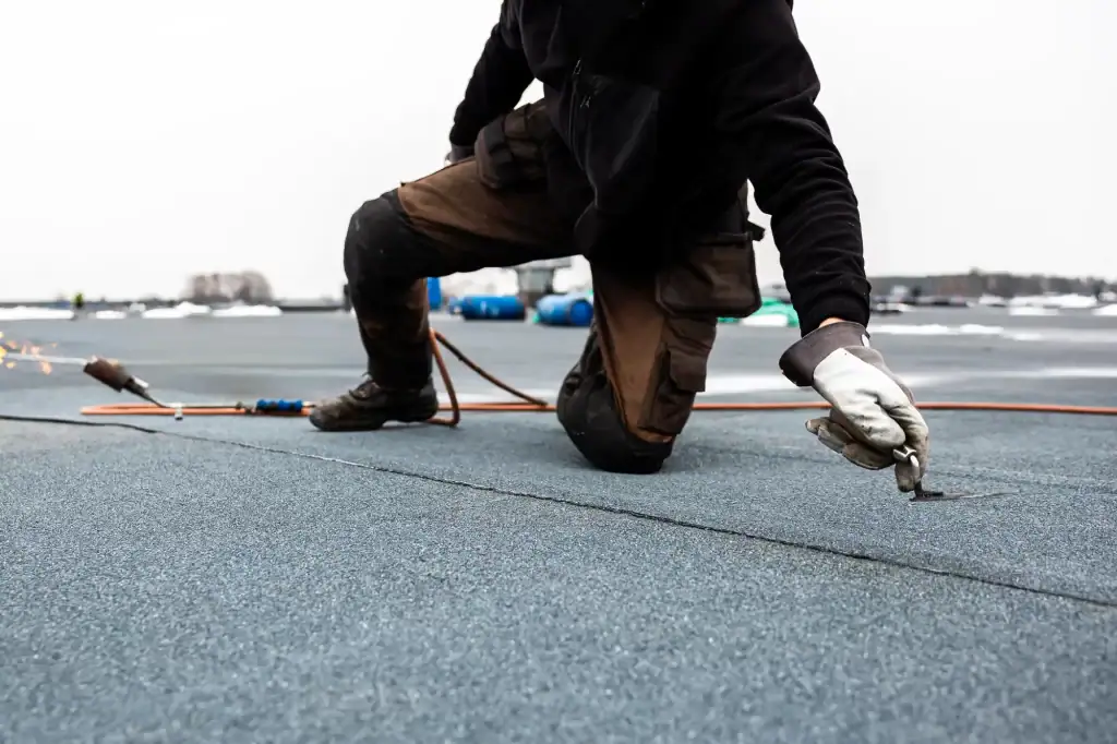 A roofer Long Island in gloves kneels on a flat roof, installing or repairing roofing material with tools and equipment visible around, on an overcast NY day.