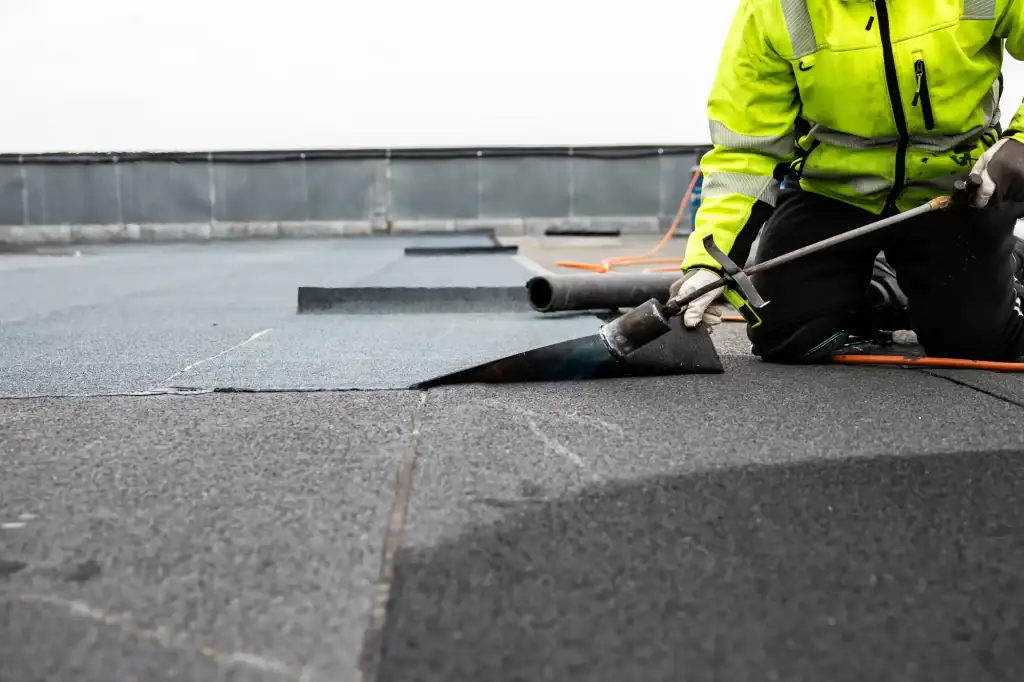 A roofer Long Island kneels on a flat rooftop, wearing a high-visibility jacket while using a torch to install roofing material. Several overlapping sheets are clearly visible across the surface.