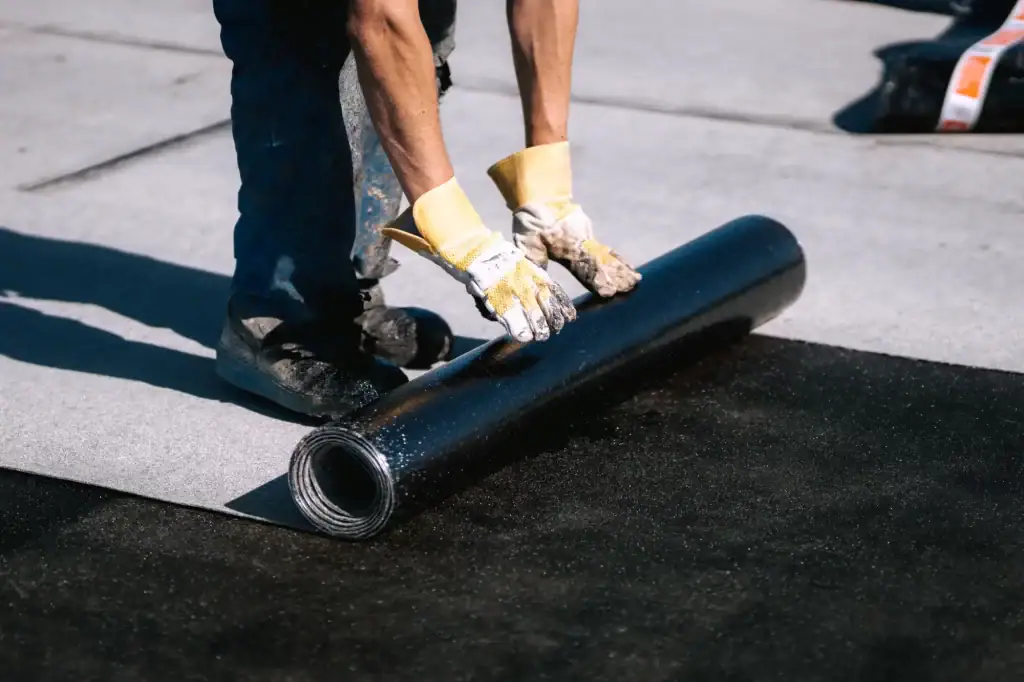 A roofing contractor Long Island unrolls a sheet of black waterproof material onto a flat roof during installation or repair, wearing protective gloves.