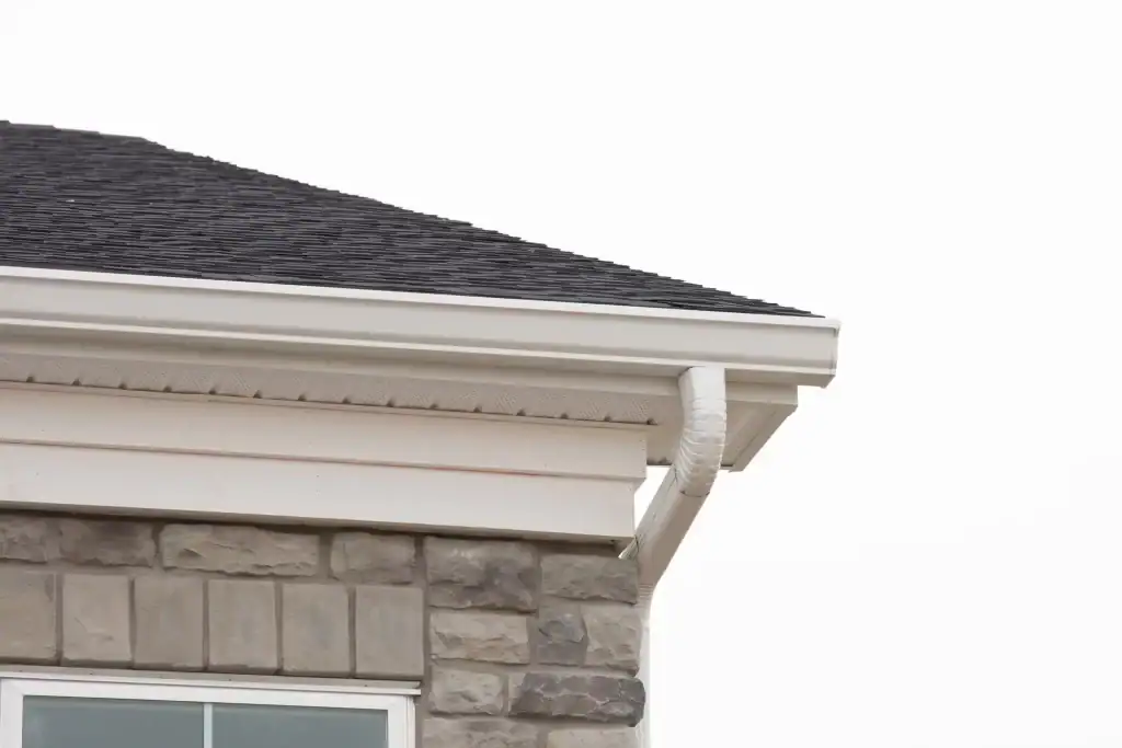 Close-up of a house roof corner with dark shingles, white gutters, a downspout, and gray stone exterior walls against a white sky—an example of quality work by a trusted roofer Long Island, NY.