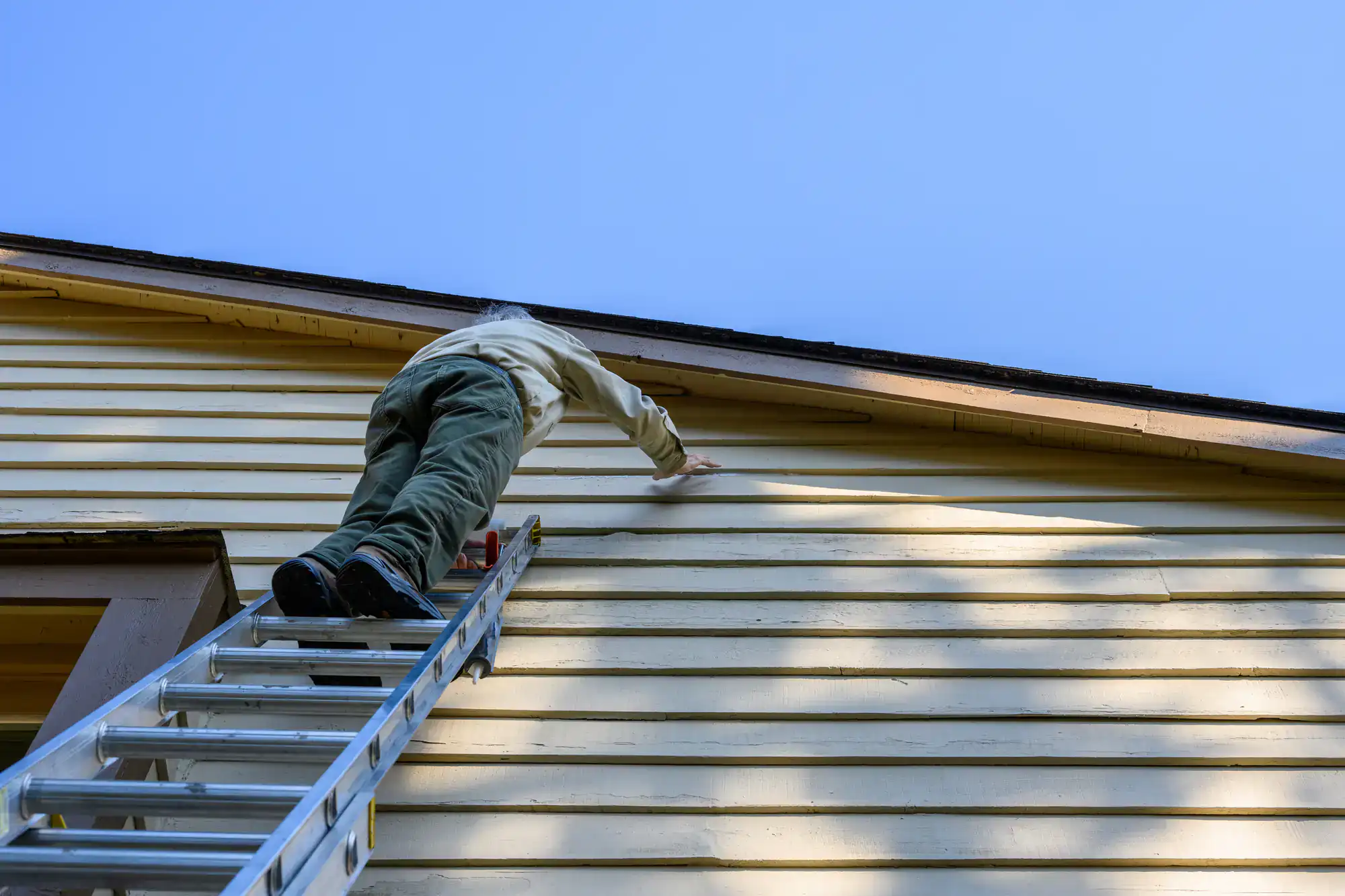 A person stands on a ladder, reaching up to touch the siding of a yellow house under a clear blue sky—capturing the view from below, just as a roofer Long Island might when assessing a NY home's exterior.