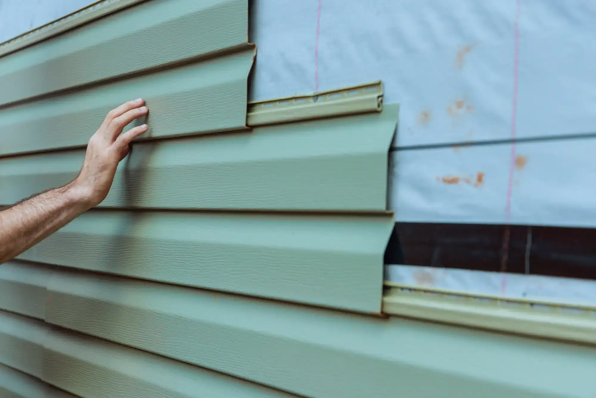 A person’s hand is installing or inspecting light green vinyl siding on the exterior wall of a house in NY, with a section of unfinished wall and weather barrier visible—precision often seen from a top roofing contractor Long Island trusts.