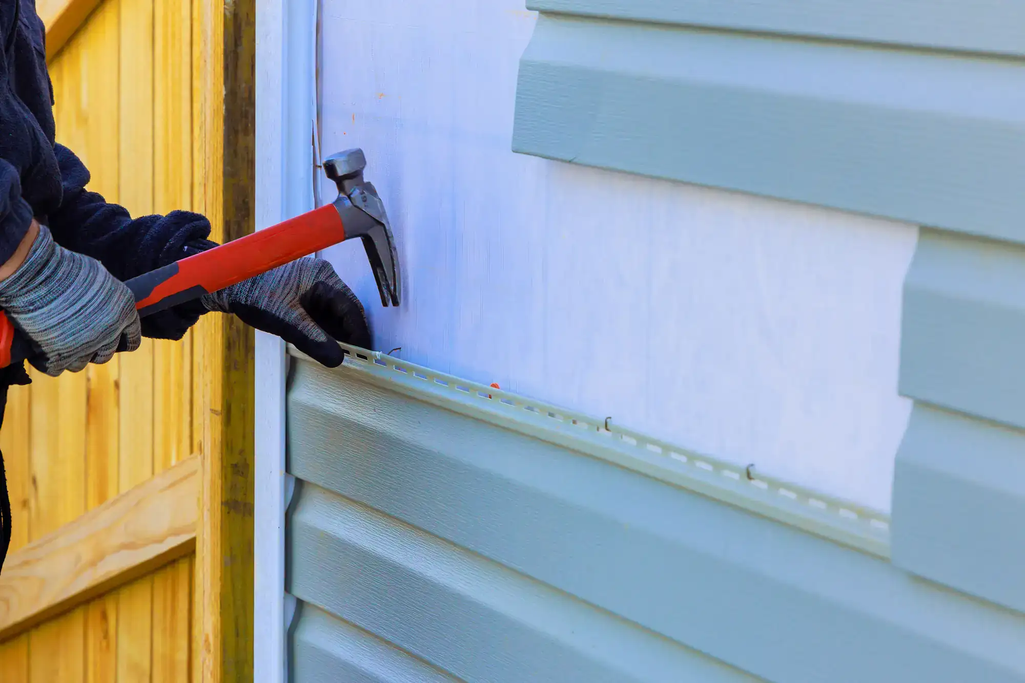 A person wearing gloves uses a hammer to install light blue vinyl siding on the exterior wall of a house near a wooden fence, showcasing the skills of a roofing contractor Long Island trusts.
