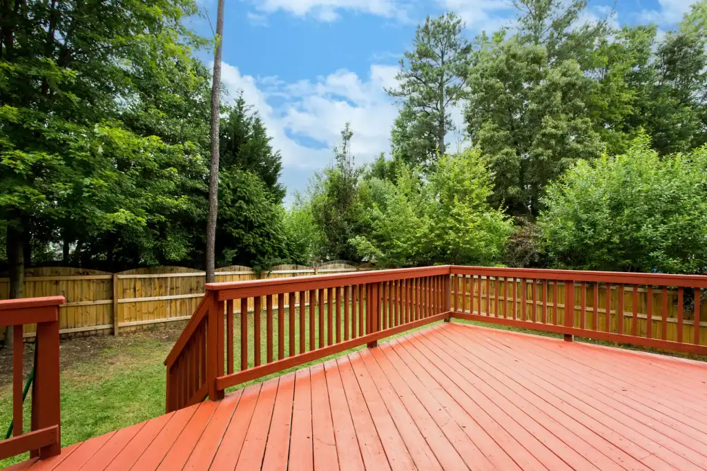 A spacious red wooden deck with railing overlooks a fenced backyard filled with green trees and grass under a partly cloudy blue sky, ideal for relaxing or meeting with your trusted roofing contractor Long Island, NY.