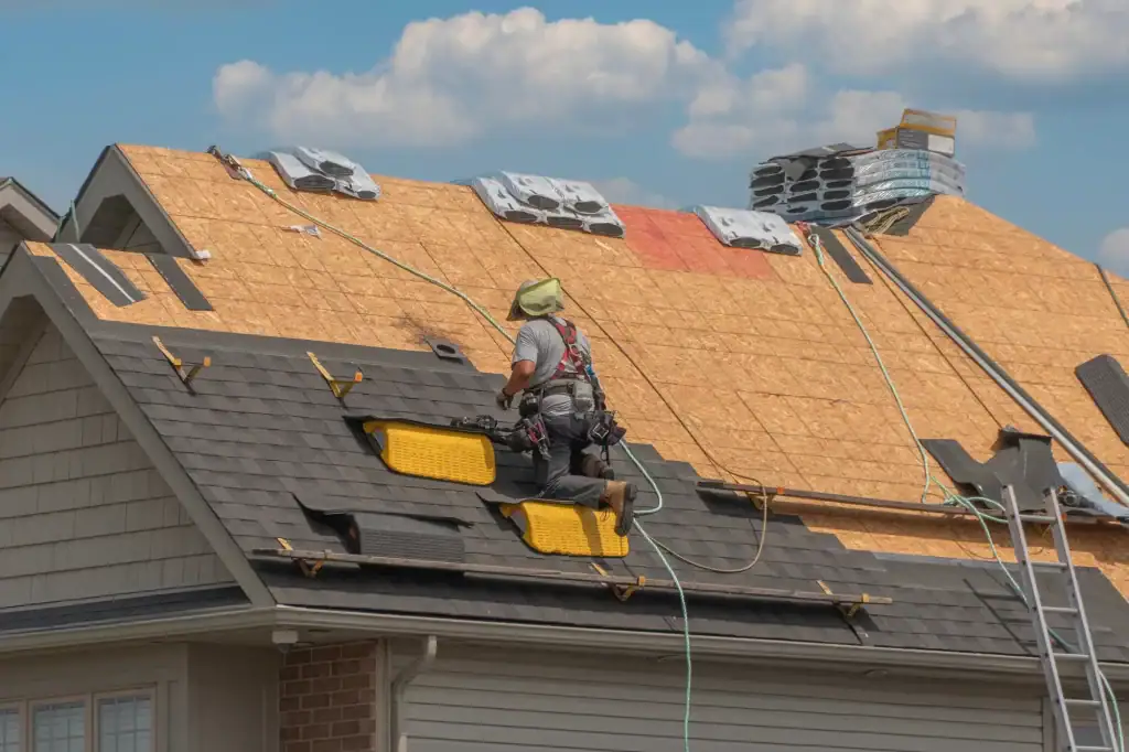 A worker in safety gear installs new shingles on a house roof under construction, showcasing the expertise of a roofer Long Island trusts; roofing materials and tools are visible under a bright NY sky with clouds in the background.