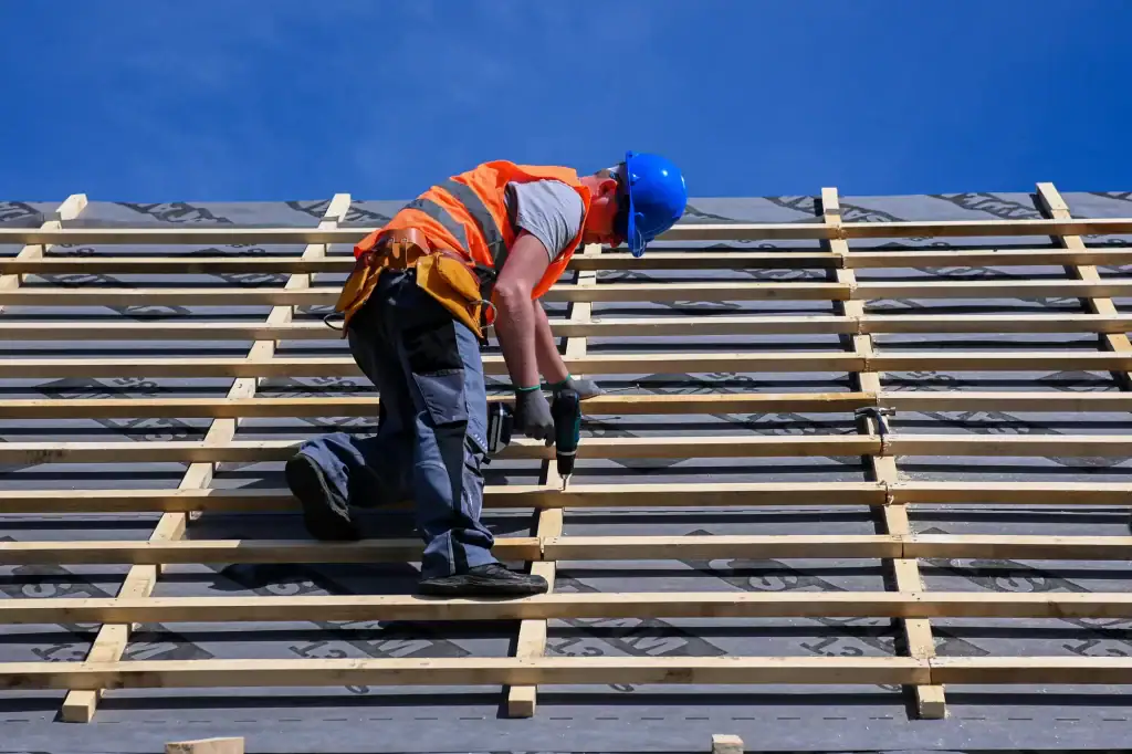 A roofing contractor Long Island kneels on a roof in a blue hard hat and orange safety vest, securing wooden battens beneath a clear blue sky.