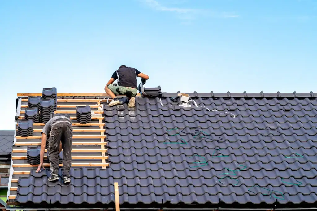 Two workers from a roofing contractor Long Island install dark roof tiles on a house under a clear blue NY sky. One arranges tiles while the other aligns them on wooden battens, with stacks of tiles and tools visible on the roof.