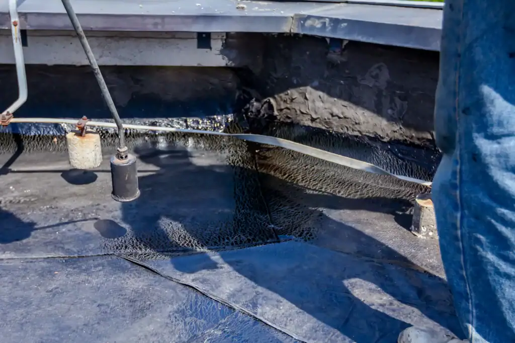 A roofing contractor Long Island applies hot bitumen with a torch to seal a flat roof. The dark, shiny surface is partially covered with roofing material. Shadows and construction tools are visible across the roof.