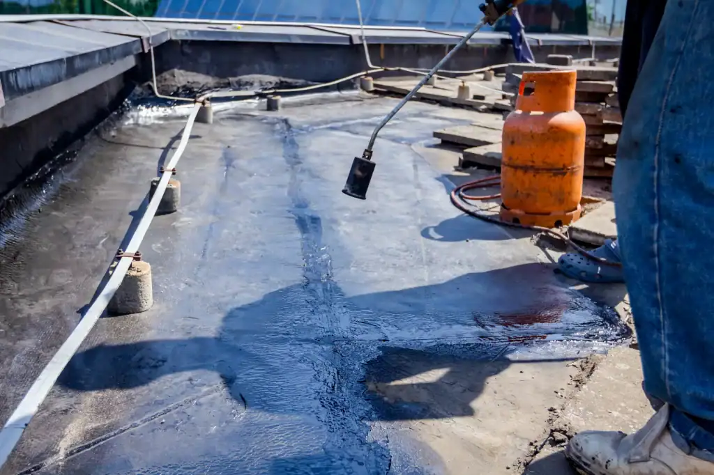 A roofer Long Island uses a propane torch to apply a waterproof membrane on a flat rooftop. An orange gas cylinder and construction materials are visible, with sunlight casting shadows on the black membrane surface.