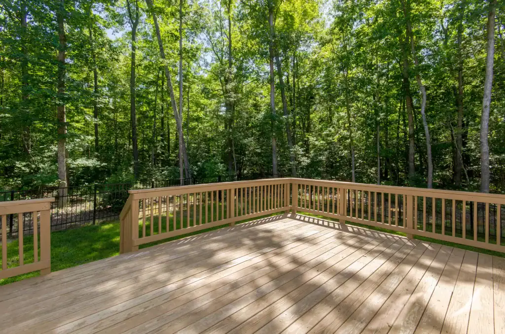 A spacious wooden deck with rails overlooks a sunny, wooded backyard filled with tall green trees and dense foliage—crafted by a trusted roofing contractor Long Island residents rely on.