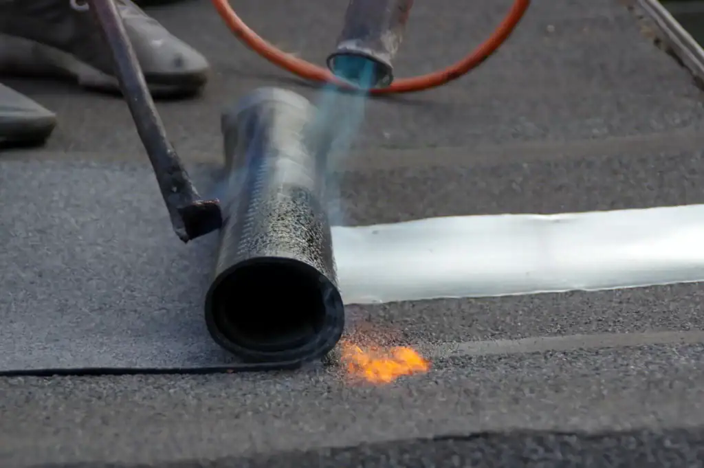 A roofer Long Island uses a blowtorch to heat and apply a roll of roofing material onto a flat roof, melting the adhesive for installation. The flame and heat are visible near the edge of the material.