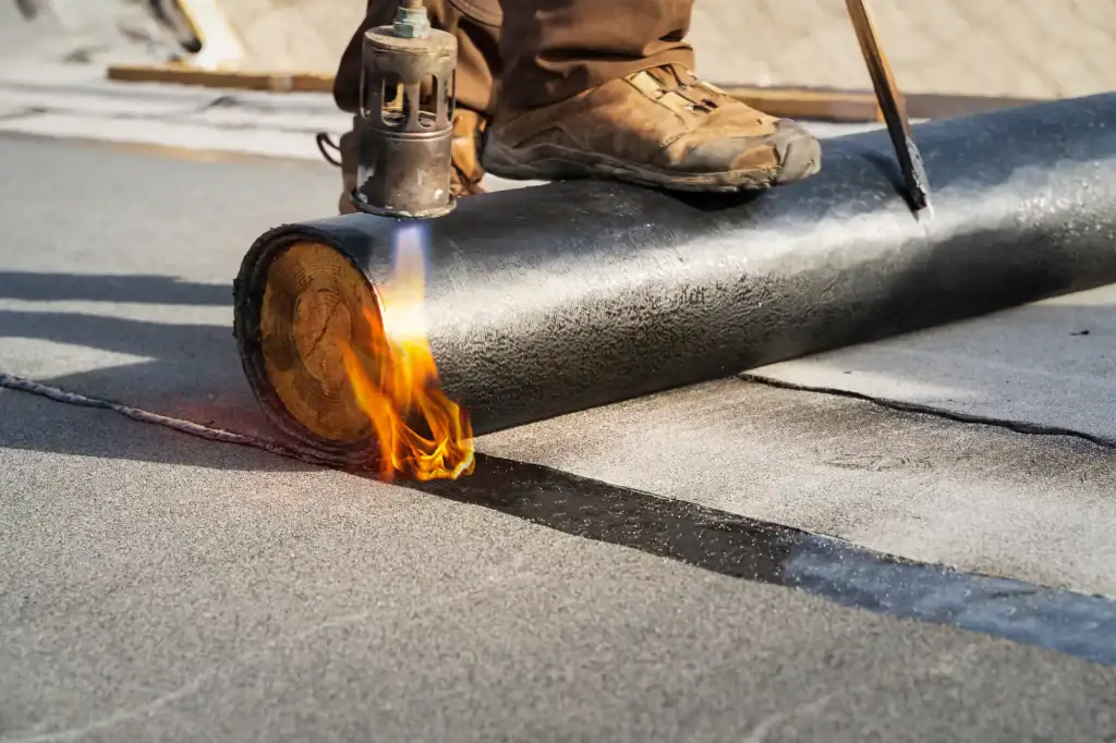 A roofer Long Island, NY uses a torch to heat and install roofing material on a flat roof, wearing brown work boots and holding a tool while flame applies heat to the material.
