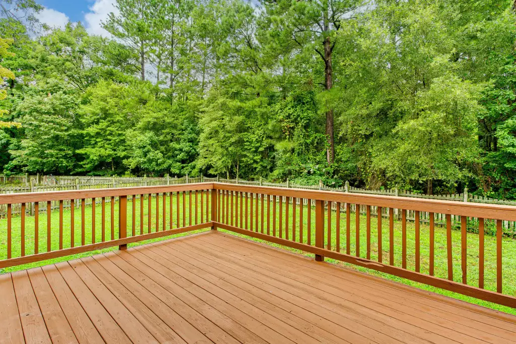 A wooden deck with a railing overlooks a grassy backyard bordered by a wooden fence and dense, green trees under a partly cloudy sky—perfect scenery for a roofing contractor Long Island project.