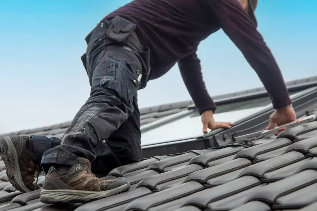 A roofer Long Island kneels on a tiled roof in NY, wearing work pants and boots, leaning forward to work on a window under a clear blue sky.