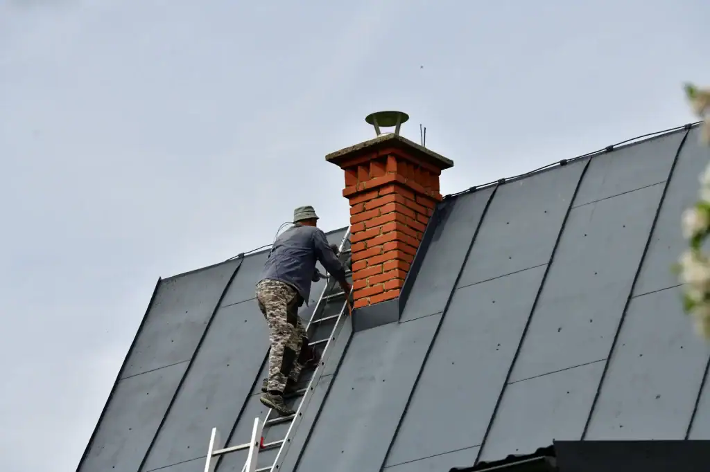 A person wearing camouflage pants and a hat climbs a ladder propped against a brick chimney on a sloped metal roof under a cloudy NY sky, resembling a seasoned roofer Long Island trusts.