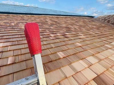 A ladder with a red padded cover rests against a newly installed wooden shingle roof under a partly cloudy sky. Part of the roof, completed by a roofer Long Island trusts, is still covered with blue underlayment.