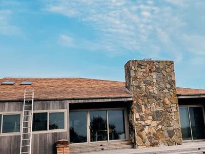 A house with large windows, a stone chimney, and a wooden roof. A roofing contractor Long Island works as a ladder leans against the roof and materials are stacked on the deck below. The sky is blue with light clouds.